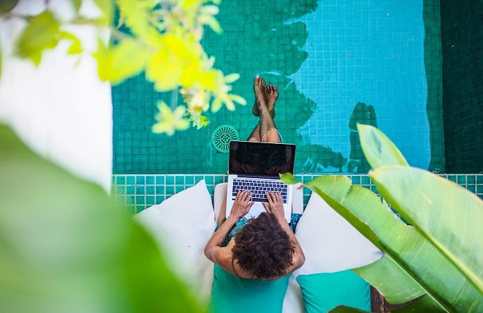 Overhead view of woman who relaxes by the side of a pool while working remotely on laptop