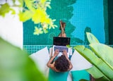 Overhead view of woman who relaxes by the side of a pool while working remotely on laptop