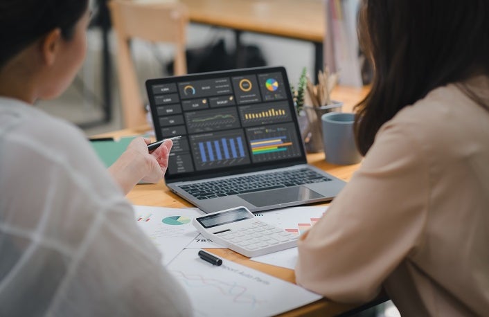 Two businesspeople or an accountant team are analyzing data charts, graphs, and a dashboard on a laptop screen in order to prepare a statistical report and discuss financial data in an office.