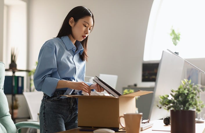 Sad unemployed Asian woman packing personal belongings in cardboard box standing at table desk, feeling stressed and depressed losing her job. Upset dismissed lady leaving office after being fired