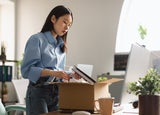 Sad unemployed Asian woman packing personal belongings in cardboard box standing at table desk, feeling stressed and depressed losing her job. Upset dismissed lady leaving office after being fired