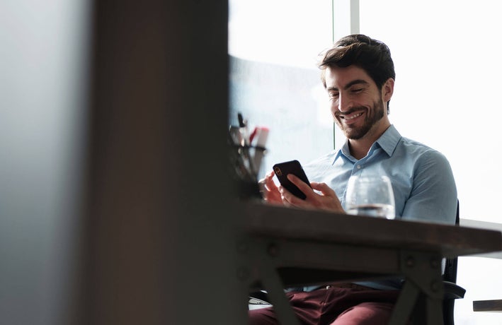 Man sitting at a desk, looking at his smartphone and smiling